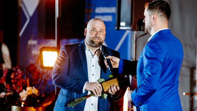 cilvēks runa mikrofonā, tur rokās ziedus un balvu, otrs cilvēks tur mikrofonu