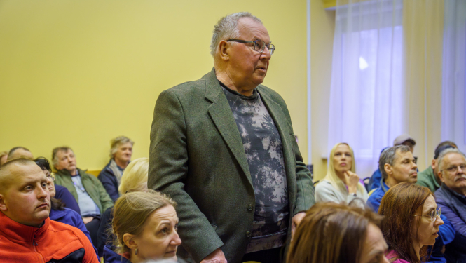 daudzi cilvēki sēž, viens vīrietis piecēlies kājās