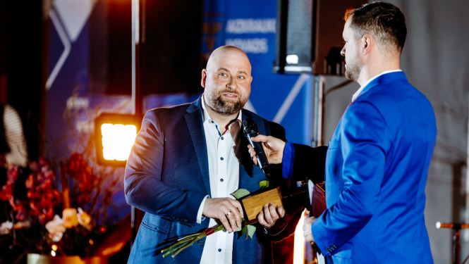 cilvēks runa mikrofonā, tur rokās ziedus un balvu, otrs cilvēks tur mikrofonu