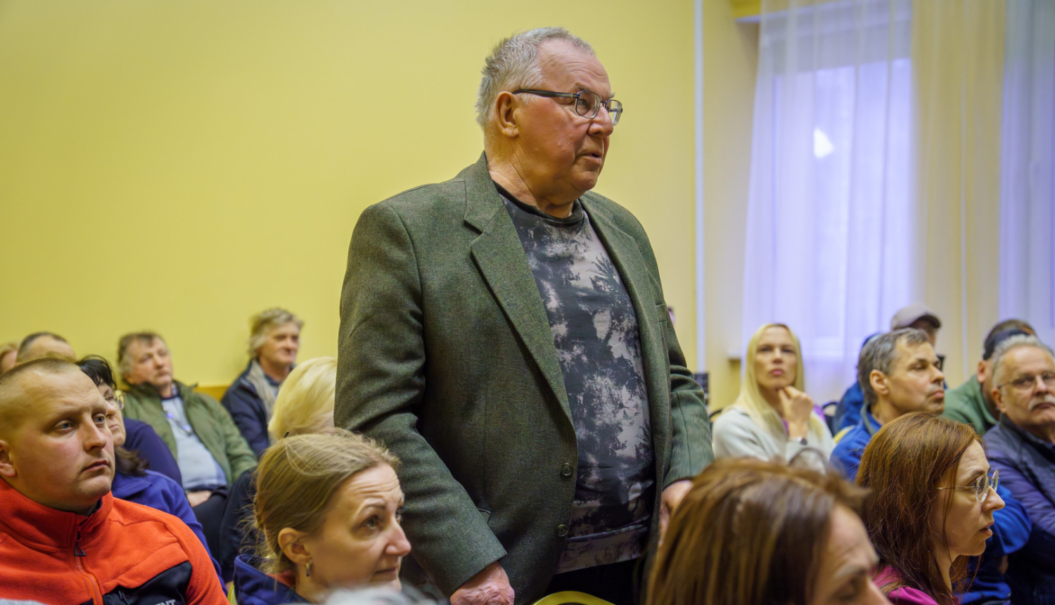 daudzi cilvēki sēž, viens vīrietis piecēlies kājās