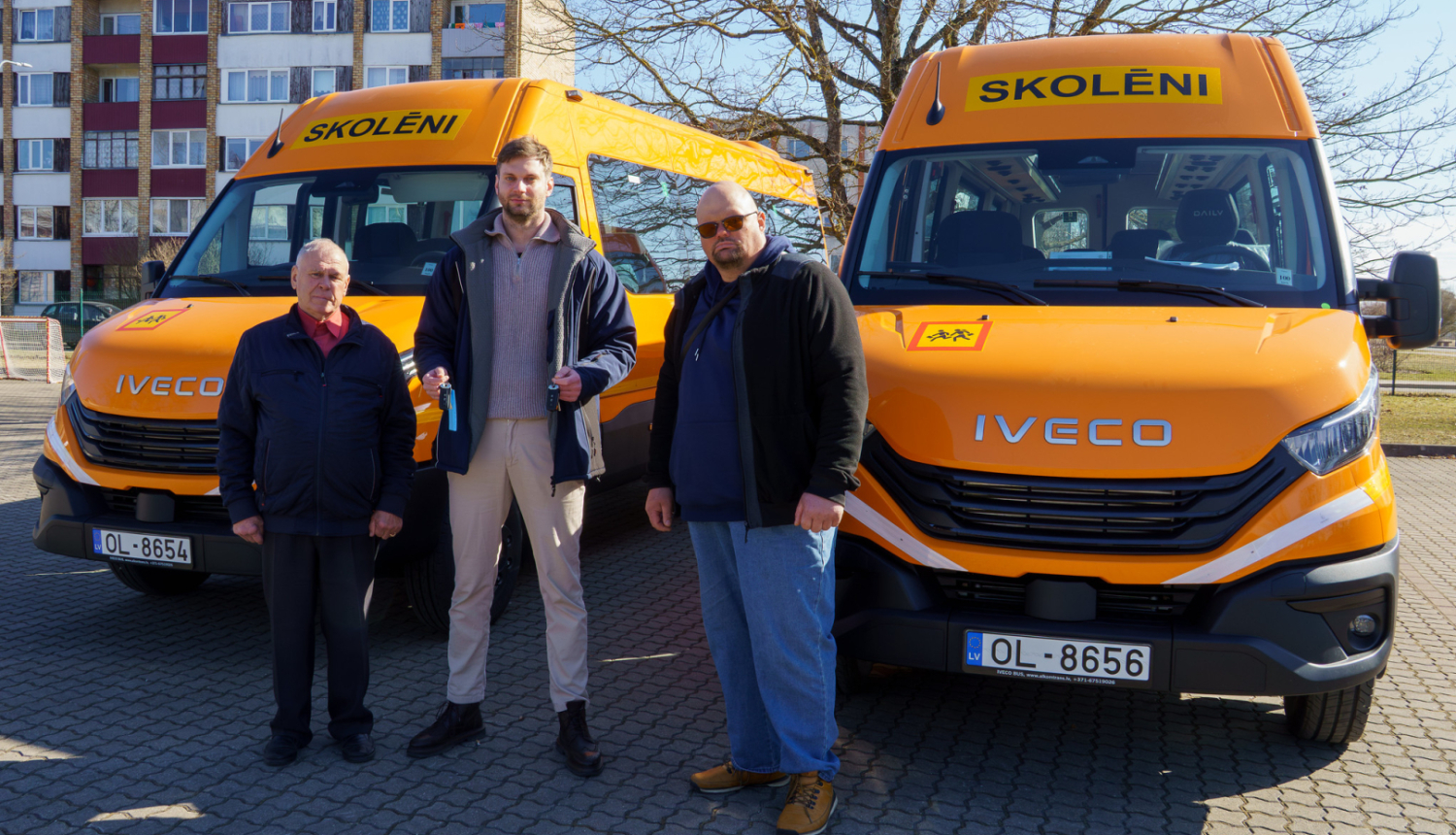 trīs vīrieši stāv pie diviem skolēnu autobusiem