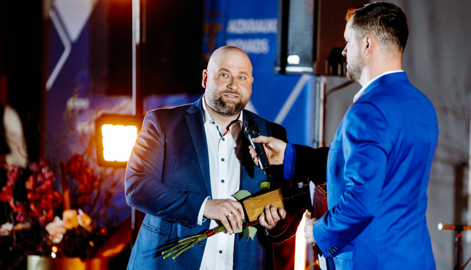 cilvēks runa mikrofonā, tur rokās ziedus un balvu, otrs cilvēks tur mikrofonu