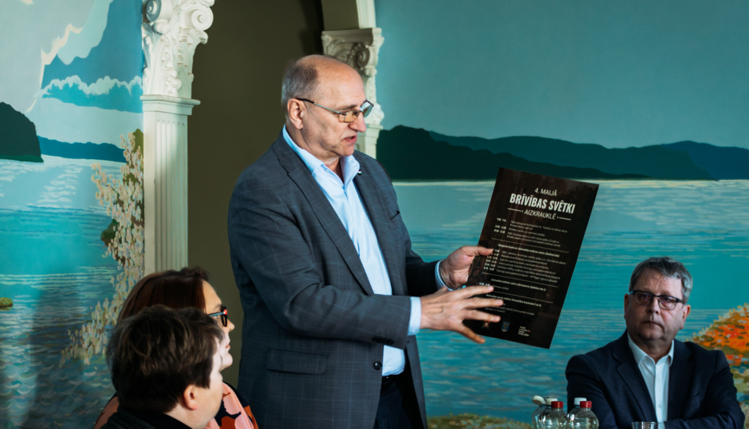 vīrietis tur rokās afišu un rāda uz to